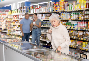 Watchful aged woman customer selecting frozen food from market fridge in superstore