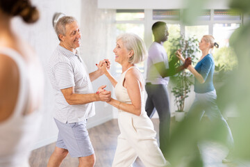 Senior man learns to dance Latin salsa dance with elderly lady. Dance studio mature clients move in background - obrazy, fototapety, plakaty