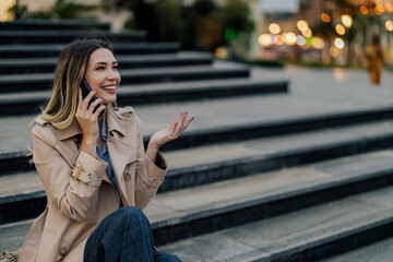 Fototapeta premium Blonde smiling woman using mobile phone while sitting on a stairs outside