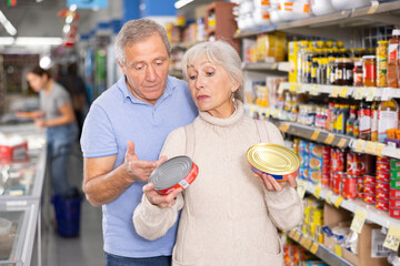 Interested senior woman and man choosing together canned food products on shelves in supermarket,...