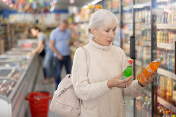 Interested elderly female customer choosing soft drinks in grocery store, looking at products on...