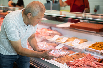 Interested elderly man looking at large assortment of fresh raw meat products in glass refrigerated...
