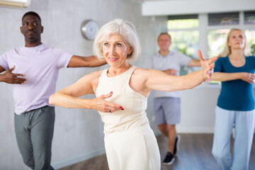 Smiling slim senior woman practicing slow foxtrot movements during beginners group class in modern...