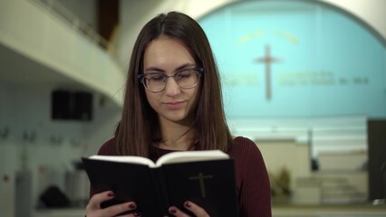 A young woman reads the Bible against the background of a cross in a church. A Protestant girl with glasses reads the Bible in church closeup.
