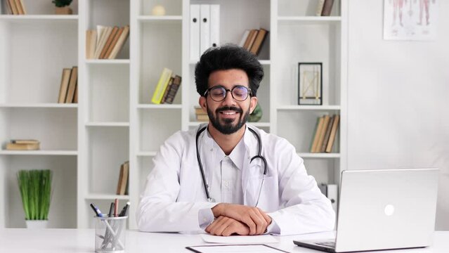 Portrait of a young Indian doctor wearing glasses smiling and looking at the camera in a white coat with a stethoscope, sitting at a table in the office. Close up view.