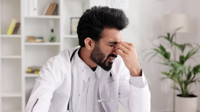 Stressed young male Indian doctor in white uniform with stethoscope looking at laptop screen, thinking about difficult solution to medical problem or feeling exhausted after hard day in hospital.