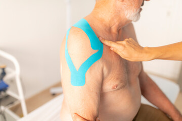 A female physiotherapist cheks the neuromuscular bandage or kinesiology tape from the shoulder of her patient, an older man - obrazy, fototapety, plakaty