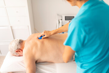 A female physiotherapist massages the back of an older man with a massage tool, a finger protector, during a manual therapy session in a clinic