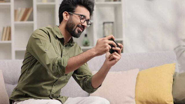 An Indian man is sitting on the sofa in his living room and playing an online game on his mobile phone, raising his hands up in victory celebration.