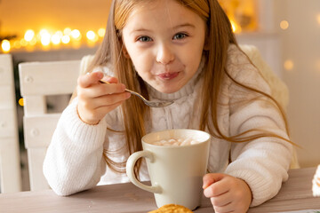 A little girl drinks cocoa with marshmallows in the kitchen at home on Christmas day. Cozy house....