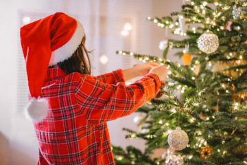  Christmas tree Decorating. Girl child in santa hat and plaid pajamas decorate the Christmas tree...