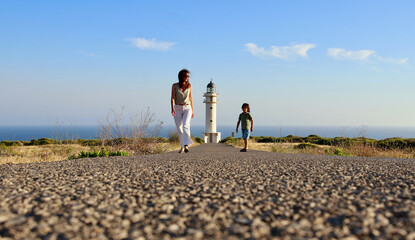 Mother and son walking towards Cap de Barberia's lighthouse, which stands beautiful on background, Formentera, Balearic Islands, Spain.