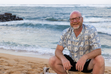 Friendly bald man with glasses and a beard wearing shorts. The middle aged guy is smiling while sitting on a log on a tropical island beach in Hawaii. He is on vacation and relaxed. 