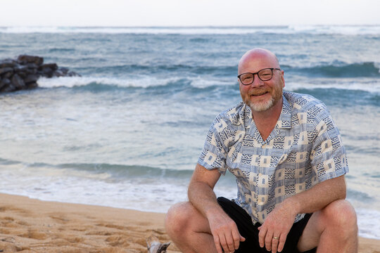 Happy bald man wearing glasses sitting at Pacific ocean beach on the Hawaiian island of Kauai while on vacation. 