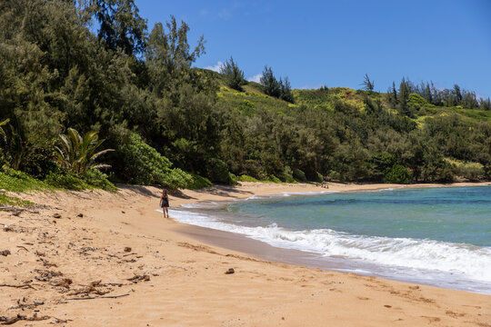 A woman walks alone on a tropical beach on the island of Kauai in Hawaii on a hot sunny, summer day next to the ocean. 