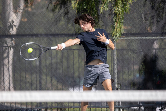 Athletic man playing tennis on a court outside. He is swinging at and hitting the ball. 