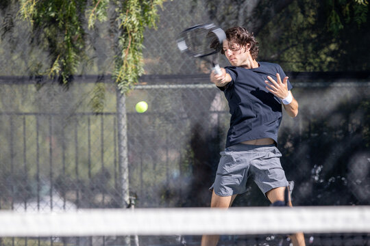 Athletic man playing tennis on a court outside. He is swinging the racket and hitting the ball. 