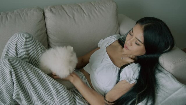 Young thai woman petting cute little white spitz dog lying on the sofa in living room with modern interior