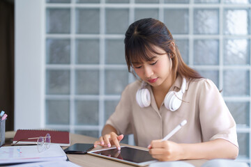 Young asian student in casual clothes wearing headphones in her neck and using tablet to writing...
