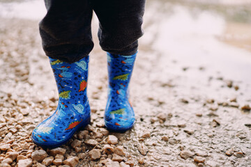 Small child in colorful rubber boots stands on gravel in a puddle. Cropped. Faceless