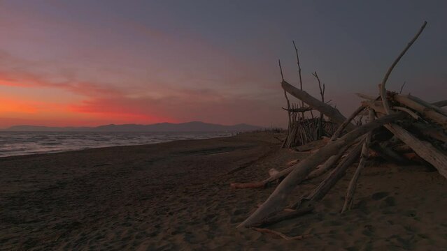 Wooden tipis on a sandy beach at sunset. Red cloud evening sky in Maremma, Tuscany, Italy. Seaside shore waves.