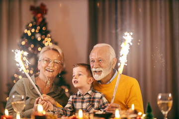 A festive grandparents sitting at home at dining table with grandson and celebrating christmas and new year's eve with fireworks. - obrazy, fototapety, plakaty