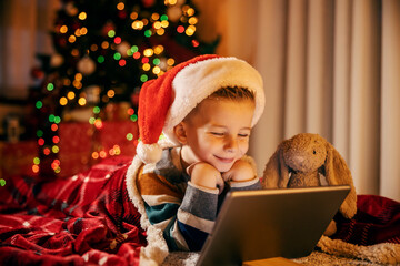 A happy boy is watching christmas cartoons at home while smiling at the tablet on christmas eve.