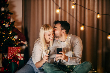 A jolly couple is kissing and using a phone while celebrating christmas and new year's eve at home with champagne.