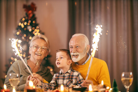 A festive grandparents sitting at home at dining table with grandson and celebrating christmas and new year's eve with fireworks.