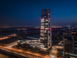 Fototapeta premium Budapest, Hungary - Aerial view of Budapest's new, illuminated skyscraper building with National Athletics Center and city skyline at background at dusk with clear blue sky