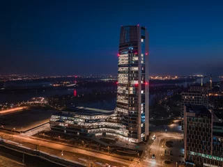 Papier Peint photo Budapest Budapest, Hungary - Aerial view of Budapest's new, illuminated skyscraper building with National Athletics Center and city skyline at background at dusk with clear blue sky