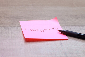 Sticky note with handwritten text I Love You with pen over wooden table. Romantic message.