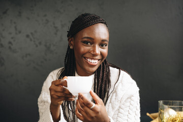 African woman in warm white sweater drinking hot coffee or tea at home