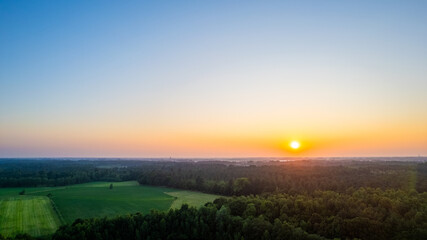 Naklejka na ściany i meble The image captures the serene beauty of sunrise over a tranquil rural landscape. The horizon is ablaze with the soft, warm hues of the early morning sun, as it rises above a dense forest and casts its