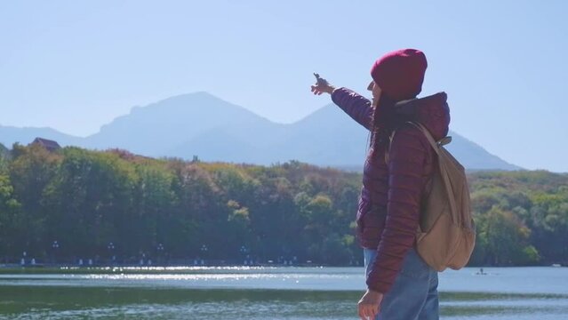 Female traveler in a burgundy jacket with a backpack on a pier near a lake in the mountains in autumn, enjoying the view of the landscape. travel to the Caucasus mountains, emotions of happiness, vaca