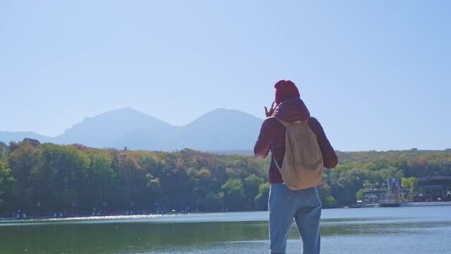 Female traveler in a burgundy jacket with a backpack on a pier near a lake in the mountains in autumn, enjoying the view of the landscape. travel to the Caucasus mountains, emotions of happiness, vaca