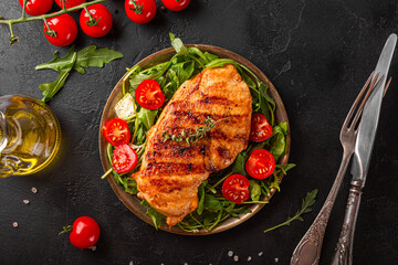 Grilled chicken breast with arugula and tomatoes on a plate, top view, black food background