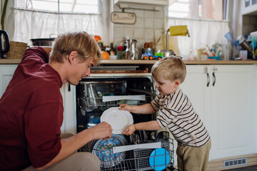 Little boy helping father to load dishwasher after breakfast. Cleaning the kitchen before leaving...