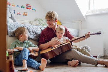 Father teaching boys to play on guitar. Boys having fun in their room with dad, playing guitar and singing together. Concept of Father's Day, and fatherly love.