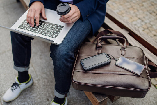 Businessman, freelancer or manager working outdoors in city park. Man with laptop on knees drinking coffee, charging his smartphone with solar phone charger. Concept of working remotely.