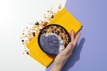 Bowl with smoothie on towel and hand on white and purple background, top view