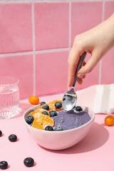 Bowl with smoothie, spoon in female hand and berries on pink background
