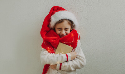 A cute teenage girl in a pullover and Santa Claus hat holding an envelope with a letter for Santa Claus against the background of a light wall. Magic Christmas atmosphere at home