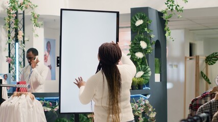 African american woman in fashion boutique using mockup augmented reality screen to look at clothes options to try on. Customer using led kiosk to visualize outfit combinations in store before buying - obrazy, fototapety, plakaty
