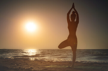 Full length rear view of a silhouette of a woman standing on one leg while practicing tree pose in yoga on a tranquil beach, taken at sunset or sunrise during summer vacation