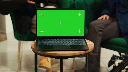 Digital device with greenscreen template placed on coffee table in lounge area at luxury alpine ski resort. People sitting on couch chatting, laptop running isolated copyspace display at hotel.