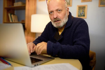 Side view of thoughtful senior novelist in elegant cardigan creating new plot of his novel, sitting in his cabinet at table in front of laptop, typing, looking at camera with thoughtful face - obrazy, fototapety, plakaty