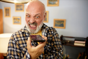 Senior man in checkered shirt yelling at smartphone. Angry furious bald bearded man recording voice message, expressing rage and displeasure, shouting with opened mouth, scolding someone - obrazy, fototapety, plakaty