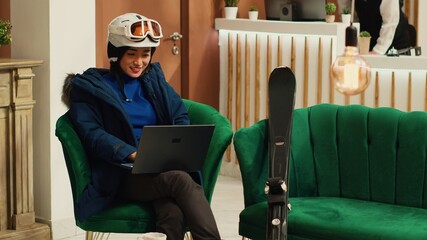 Tourist reading reviews on laptop at luxury hotel, sitting in lounge area with skiing equipment and accessories. Asian woman working remote on pc before practicing extreme winter sport in the cold.