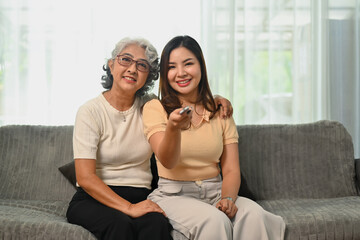 Happy retired woman relaxing on sofa with grown up daughter and watching tv on weekend